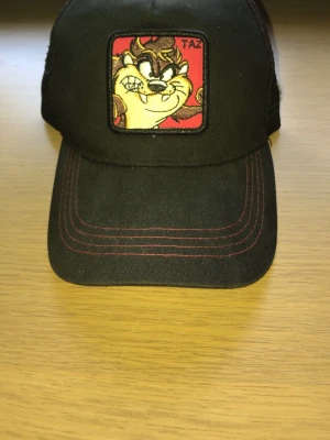 Svart keps med Taz-brodyr - Säljer en svart keps med röd kontrastsöm och broderad Taz från Looney Tunes framtill. Kepsen har justerbar plastrem bak och meshmaterial på sidorna för extra ventilation. Perfekt för dig som gillar tecknade klassiker och vill sticka ut.