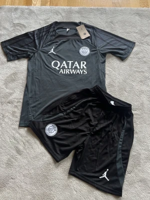 PSG träningsset i svart - Snyggt svart träningsset med t-shirt och shorts från PSG. Setet har vita loggor, bland annat Jordan och PSG-emblem, samt Qatar Airways-tryck på tröjan. Materialet är lätt och andas, perfekt för träning eller fotboll. Elastisk midja på shortsen för skön passform.
