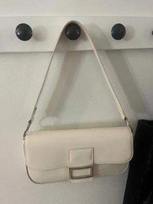 Krämvit axelväska med silverdetalj -  axelväska i krämvitt konstläder med rektangulär form och bred axelrem. Väskan har en stor silverfärgad spännesdetalj på framsidan och minimalistisk design utan synligt märke. 