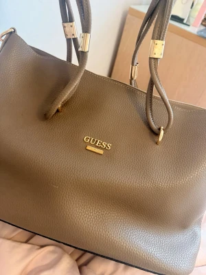 Beige handväska från Guess - Snygg beige handväska från Guess i strukturerat konstläder med guldfärgade detaljer och dubbla axelband. Väskan har en stilren design med Guess-logga framtill och dragkedja upptill. Perfekt för dig som vill ha en trendig och rymlig väska.