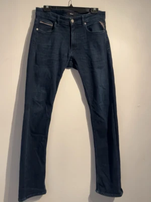 Mörkblå replay anbass jeans - Snygga mörkblå slim fit jeans med klassisk femficksdesign och raka ben från replay. Jeansen har silverfärgade knappar och detaljer samt en patch bak i midjan. Perfekta för en trendig och avslappnad stil. Modell anbass