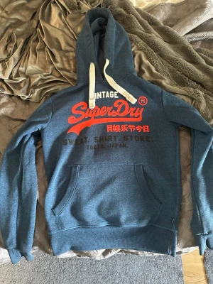 Blå Superdry hoodie  - Säljer en blå hoodie från Superdry med stort rött och svart tryck på bröstet och vita snören i huvan. Klassisk känguruficka framtill och ribbade muddar vid ärmar och midja. Perfekt för dig som gillar streetwear och vill ha en snygg och bekväm look. 9/10