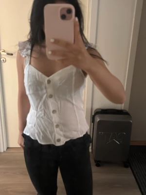 Vit korsettopp med volangdetaljer - Säljer en söt vit korsettopp med volangprydda axelband och knappar framtill. Toppen har en figurnära passform och v-ringad hals. Perfekt för dig som vill ha en romantisk och trendig look. Säljer den eftersom den är för stor för mig och inte då kommer till användning 