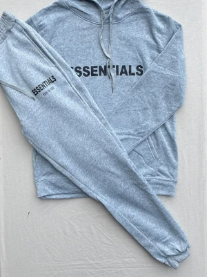 Grått Essentials loungewear-set - Mysigt grått loungewear-set från Essentials Fear of God med hoodie och matchande byxor. Setet har tryckt logga på både tröjan och byxan, dragsko i midjan och huva samt muddar vid ärmslut och benslut. Perfekt för chill dagar hemma.