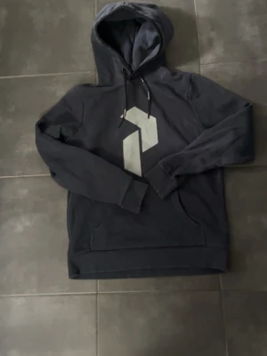 Marinblå peak performance hoodie med stor logga - Snygg marinblå hoodie från Peak Performance med stor grå logga på bröstet och justerbar huva med snören. Tillverkad i mjukt material och har en klassisk känguruficka framtill. Perfekt för en avslappnad och sportig stil.