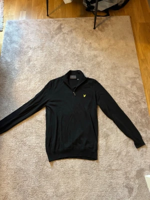 Svart stickad tröja med dragkedja - Svart stickad tröja med halv dragkedja och gul broderad logga på bröstet. Perfekt för lager-på-lager och enkel att matcha med olika outfits.