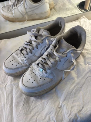 Vita Nike Air Force 1  - Klassiska vita Nike Air Force 1 sneakers med lågt skaft och perforerad tå. Skorna har en chunky sula och ikonisk Swoosh på sidorna. Tillverkade i läder med textilfoder och snörning framtill. Perfekta för en clean och stilren streetlook.