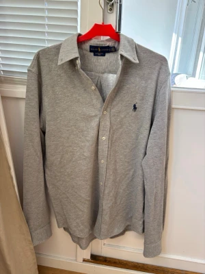 Grå långärmad skjorta från Ralph Lauren - Säljer en stilren grå skjorta från Ralph Lauren i mjuk bomull. Skjortan har klassisk krage, knappar framtill och den ikoniska mörkblå loggan broderad på bröstet. Perfekt för dig som gillar enkel och tidlös stil.