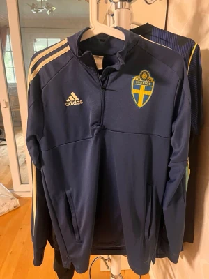 Blå svensk träningsjacka Adidas - Mörkblå träningsjacka från Adidas med Sveriges landslagsemblem på bröstet och vita ränder på axlarna. Jackan har halv dragkedja, ståkrage och två sidofickor. Tillverkad i polyester, perfekt för träning eller uppvärmning.
