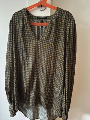Mönstrad blus med V-ringning - Snygg blus med småmönstrat tryck i svart och beige. Blusen har V-ringning och långa ärmar med lätt puff vid axlarna. Tillverkad i ett tunt, mjukt tyg som faller fint. Perfekt för dig som gillar en avslappnad men ändå stilren look.