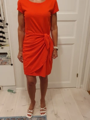 Röd kortklänning med knyt i midjan - Snygg röd kortklänning med rund halsringning och korta ärmar. Klänningen har ett omlottliknande fall framtill och ett dekorativt knyt i midjan som ger en cool och avslappnad look. Tillverkad i mjukt bomullstyg som känns skönt mot huden.