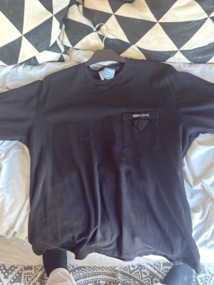 Svart t-shirt från Prada Milano - Snygg svart t-shirt från Prada Milano med klassisk passform. T-shirten har en bröstficka med ikonisk Prada-logga och en liten tygdetalj i form av en triangel. Perfekt till jeans eller shorts för en clean och stilren look.