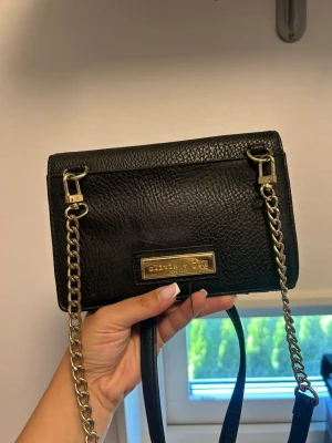 Svart handväska med guldkedja - Snygg svart handväska från Olivia + Joy i strukturerat konstläder. Väskan har guldfärgade detaljer, kedjerem och en metallplatta framtill. Baksidan har en dragkedjeficka och dekorativa nitar. Perfekt storlek för det viktigaste.