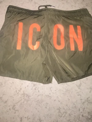 Gröna badshorts med orange text - Snygga gröna badshorts med stora orange bokstäver 'ICON' på baksidan och liten Dsquared2-logga framtill. Shortsen har snörning i midjan och är perfekta för sommaren. Materialet känns lätt och snabbtorkande.