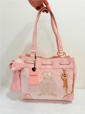 Rosa handväska med rosett Juicy Couture - Söt rosa handväska från Juicy Couture i mjuk velour med detaljer i guld, stor rosett på sidan och dekorativ kedja med nyckel. Väskan har dubbla handtag, dragsko upptill och ett broderat emblem framtill. Perfekt för dig som gillar en girly och trendig stil.