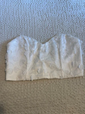 Vit blommig topp - Supersöt vit topp med blommor och broderade detaljer. Toppen är axelbandslös och har knäppning bak. Perfekt till sommaren och ger en romantisk vibe till din outfit.