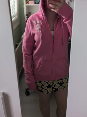 Rosa hoodie med dragkedja - Säljer en rosa hoodie från Abercrombie & Fitch med dragkedja framtill och två fickor. Tryck i vitt på bröstet med texten 'A&F NY 1892'. Mjuk bomullskänsla och huva med dragsko. Perfekt för chill dagar eller när du vill ha en färgklick i garderoben. Står L men skulle säga att den passar M
