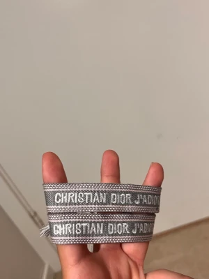 Grått armband Christian Dior - Snyggt grått tygarmband från Christian Dior med texten 'CHRISTIAN DIOR J'ADIOR' i vitt. Armbandet har en vävd struktur och justerbar knytning med tofsar i ändarna. Perfekt accessoar för att lyfta din outfit.