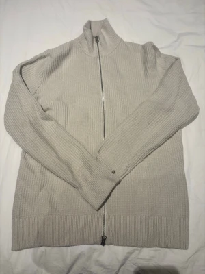 Beige stickad ziptröja Samsøe Samsøe - M passar S bättre Snygg beige stickad ziptröja från Samsøe Samsøe med hög krage och ribbad struktur. Tröjan har dragkedja hela vägen och långa ärmar. Perfekt för lager på lager och enkel att styla till olika outfits.