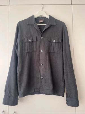 Oscar Jacobson overshirt - Stilren mörkgrå overshirt med raka linjer och två bröstfickor med lock och knapp. Skjortan har klassisk krage, knäppning framtill och långa ärmar med manschett. Tillverkad i ett mjukt tyg som ger en avslappnad look. Den är använd men väl omhändertagen😊