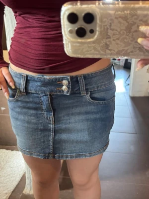Blå lågmidjad jeanskjol med dubbla knappar - Säljer en klassisk blå lågmidjad jeanskjol med dubbla silverknappar i midjan och femficksmodell. Kjolen har en kort passform och är tillverkad i slitstarkt denimtyg. Perfekt att matcha med en croppad topp eller hoodie. Skulle säga att den sitter som en S!