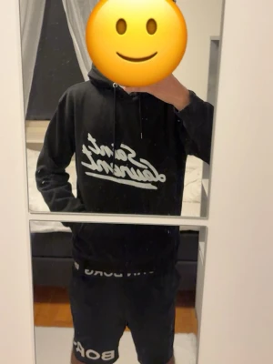 Svart Saint Laurent hoodie - Säljer en svart hoodie från Saint Laurent med vit logga i snyggt skript på bröstet. Klassisk huva med dragsko och ribbade muddar vid ärmar och nederkant. Perfekt för en chill och stilren look.