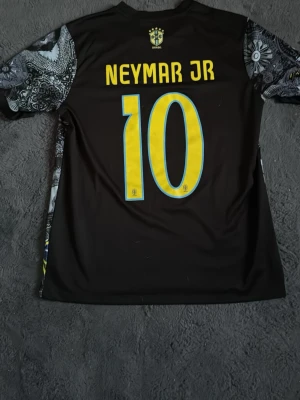 Svart Brasil Neymar JR tröja - Säljer en svart Brasilien fotbollströja från Nike med Neymar JR och nummer 10 i gult på ryggen. Framsidan har ett stort tryck av Kristusstatyn och detaljerade grafiska mönster i grått och vitt. Korta ärmar och rund hals. Perfekt för dig som älskar fotboll och vill sticka ut.