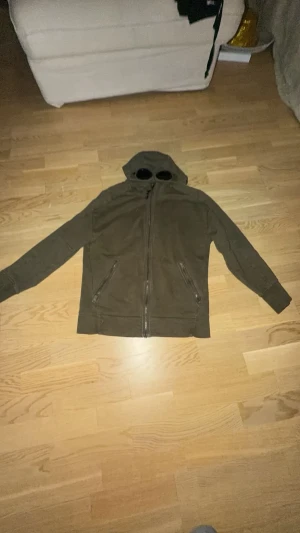 Olivgrön hoodie med glasögon i huvan - Säljer en olivgrön hoodie med dragkedja och två fickor framtill. Huvan har inbyggda glasögon vilket ger en unik och cool look. Perfekt för dig som vill sticka ut lite extra. Tillverkad i mjukt material. Den har tappat lite färg vid kedjan men bara lite. Man får även med ett par snören.