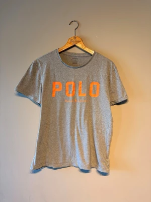 Grå POLO t-shirt från Ralph Lauren - Snygg grå t-shirt från Ralph Lauren med stor orange POLO-text och logga framtill. Klassisk rund halsringning och korta ärmar. Perfekt till jeans eller shorts för en avslappnad och trendig look.