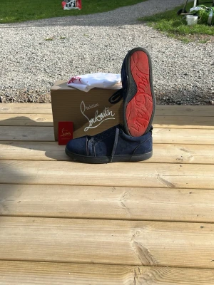Mörkblå sneakers från Christian Louboutin - Snygga mörkblå sneakers från Christian Louboutin med klassisk röd sula och svart ovandel i mocka. Skorna har låga snitt och svarta skosnören. Perfekta för dig som vill sticka ut med exklusiva detaljer och unik design.