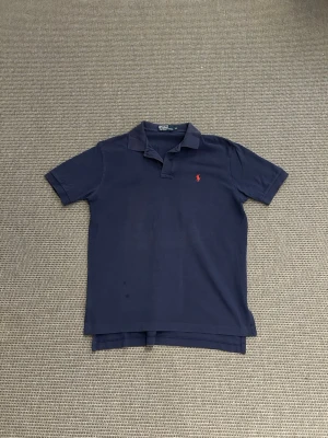 Ralph lauren piké - Klassisk marinblå pikétröja från Ralph Lauren med korta ärmar och den ikoniska röda logotypen broderad på bröstet. Tillverkad i mjuk bomull och har en stilren krage med knappslå. Perfekt till jeans eller shorts för en clean look.