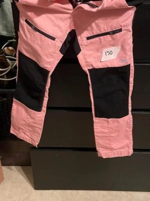 Rosa och svarta täckbyxor - Säljer ett par rosa täckbyxor med svarta förstärkningar på knän och bak, samt dragkedjefickor framtill. Perfekta för kalla dagar och utomhusaktiviteter. Byxorna har en sportig look och är tillverkade i slitstarkt material.