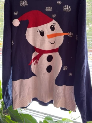 Mörkblå jultröja med snögubbe - Rolig mörkblå stickad jultröja med en stor snögubbe på framsidan, röd tomteluva och halsduk samt vita snöflingor som dekoration. Tröjan har rund hals och ribbade muddar. Perfekt för julmys och vinterhäng!