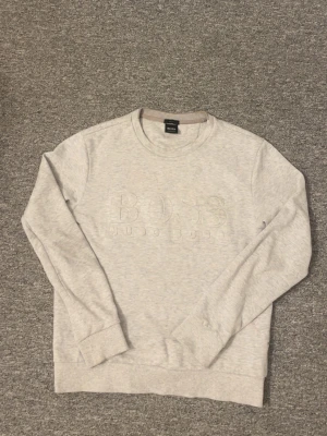 Ljusgrå sweatshirt från BOSS - Snygg ljusgrå sweatshirt från BOSS med diskret broderad logga på bröstet. Tröjan har rund halsringning, långa ärmar och ribbade muddar vid ärmslut och nederkant. Perfekt för en clean och stilren look.