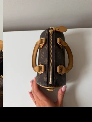Louis vuitton nano speedy bag - Snygg liten handväska från Louis Vuitton i klassiskt brunt monogrammönster. Väskan har två korta handtag i ljust läder och en justerbar axelrem. Guldiga metalldetaljer och dragkedja upptill. Perfekt för dig som gillar lyxiga accessoarer.