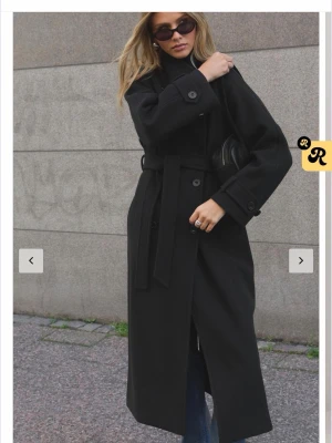 Svart lång trenchcoat med knytskärp - Stilren svart trenchcoat i lång modell med knytskärp i midjan och stora knappar ( det följer ej med knytskärp tyvärr)!!!. Klassisk krage och raka ärmar med knappdetaljer vid handlederna. Perfekt för en trendig och tidlös look. Passar till lager-på-lager och funkar året runt.
