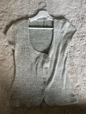 Grå topp med knappar från Zara - Säljer en ljusgrå/grön topp från Zara med korta ärmar och djup v-ringning. Toppen har fyra stora knappar framtill och är figurnära i modellen. Perfekt att styla med jeans eller kjol för en trendig look.