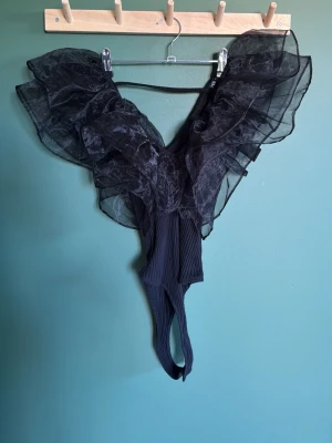 Svart body med volanger från Zara stl S - Snygg svart body från Zara med stora, transparenta volanger i mesh över axlarna och djup urringning. Kroppsdelen är ribbad och stretchig för en bekväm passform. Perfekt för dig som vill sticka ut med en unik och trendig look.