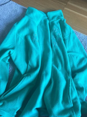 Turkos fleecejacka med stjärnor - Mysig turkos fleecejacka med dragkedja och broderade stjärnor på bröstet. Perfekt för kyliga dagar och enkel att slänga på sig. Jackan har hög krage och mjukt material som känns skönt mot huden.