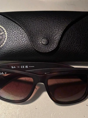 Mörkbruna/Havanna Ray-Ban solglasögon - Säljer ett par klassiska svarta Ray-Ban solglasögon med mörka glas och bred båge. Modellen Justin har en tidlös design och kommer med original svart fodral i skinnimitation. Perfekta för att höja din stil i solen. Skriv om ni är intresserade! Tyvärr fick dessa sig en smäll under semestern så fodralet släppte på högra sidan och att bågen på höger sticker ut en aningen och dermed priser. Nypris 1400kr. Självklart är det inget annat fel på dem och dem funkar precis som nya! 