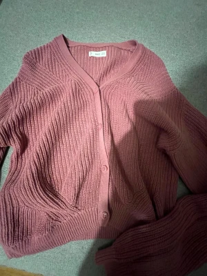 Rosa stickad kofta med knappar - Mysig stickad kofta i dammigt rosa med v-ringning och knäppning framtill. Koftan har ribbstickade muddar och ett snyggt strukturmönster i garnet. Perfekt att slänga över en t-shirt eller topp för extra värme.