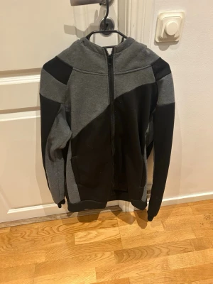Svart och grå hoodie med dragkedja - Säljer en stilren hoodie i svart och grått med hel dragkedja framtill. Hoodien har en stor huva och är tillverkad i mjukt sweatshirtmaterial. Snygg färgblock-design och långa ärmar. Perfekt för chill dagar eller när du vill ha en avslappnad look.