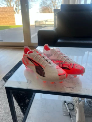 Vita och neonröda Puma fotbollsskor - Säljer ett par lätta fotbollsskor från Puma i vitt och neonrött med svarta detaljer. Skorna har låg profil, snörning och en sula med fasta dobbar för bra grepp på planen. Snygg design med ULTRA-tryck på hälen och meshmaterial för ventilation.