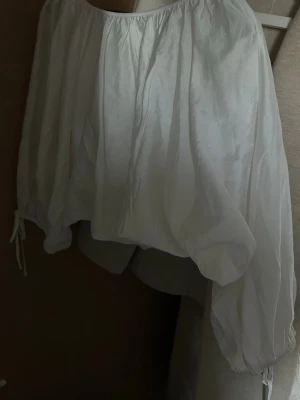 Vit offshoulder blus med ballongärm - Säljer en vit offshoulder blus med bred resår upptill och voluminösa ballongärmar som avslutas med knytband. Tunt, lätt tyg som ger en luftig känsla och trendig look. Perfekt att styla med jeans eller kjol för en cool vibe.