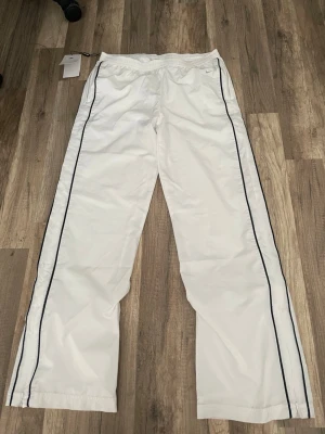 Vita mjukisbyxor från Nike - Snygga vita mjukisbyxor från Nike med svarta ränder längs benen. Byxorna har resår i midjan, dragkedjeförsedda fickor och är tillverkade i ett lätt, sportigt material. Perfekta för en avslappnad och trendig look. Användt 1 gång men har tyvärr ingen original förpackning med dem