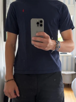 Mörkblå t-shirt från Polo Ralph Lauren - Säljer en mörkblå t-shirt från Polo Ralph Lauren med klassisk röd logga broderad på bröstet. T-shirten har rund hals och korta ärmar, perfekt basic-plagg till garderoben. Tillverkad i mjuk bomull för skön känsla. Storlek 14-16