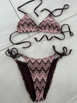 Rosa och brun zigzag-mönstrad bikini - Säljer en snygg bikini med zigzag-mönster i rosa, brun och vit. Överdelen är trekantsmodell med knyt i nacke och rygg, och underdelen har knyt i sidorna. Vackra detaljer i guld. Perfekt för stranden eller poolen i sommar!