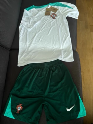 Fotbolls-dress män, Portugal - Säljer ett Portugal dress med vit tröja och gröna detaljer samt gröna shorts. Tröjan har Portugal-emblemet på bröstet och är tillverkad i Dri-FIT material som andas. Perfekt för träning eller match!