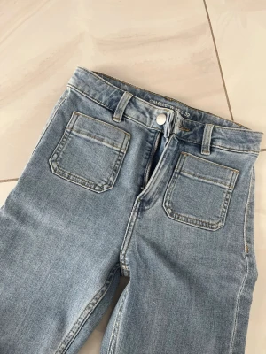 Ljusblå utsvängda jeans med fickor - Säljer ett par ljusblå jeans med utsvängda ben och två stora fyrkantiga fickor framtill. Jeansen har hög midja, bälteshällor och klassisk dragkedja med knapp. Perfekta för dig som gillar retrostil och bekväma passformer. Oanvända.