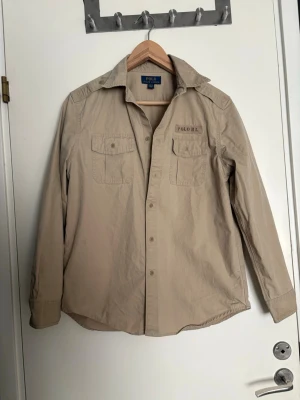 Beige skjorta/overshirt från Polo Ralph Lauren - Snygg beige skjorta/overshirt från Polo Ralph Lauren med klassisk krage och bröstficka med knapp. Skjortan har en patch med texten 'POLO RL.' ovanför fickan och är tillverkad i bomull. Perfekt för en clean och stilren look.